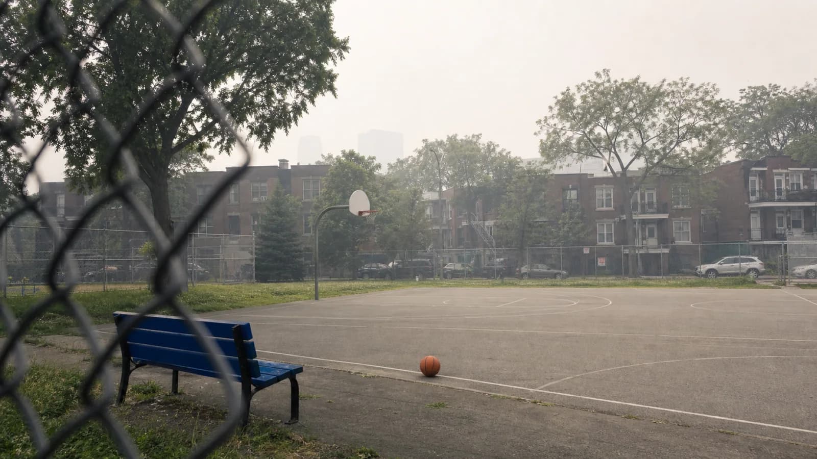 An empty Montréal neighbourhood basketball court sits under pale wildfire haze, with a blue bench and ball suggesting paused outdoor plans.