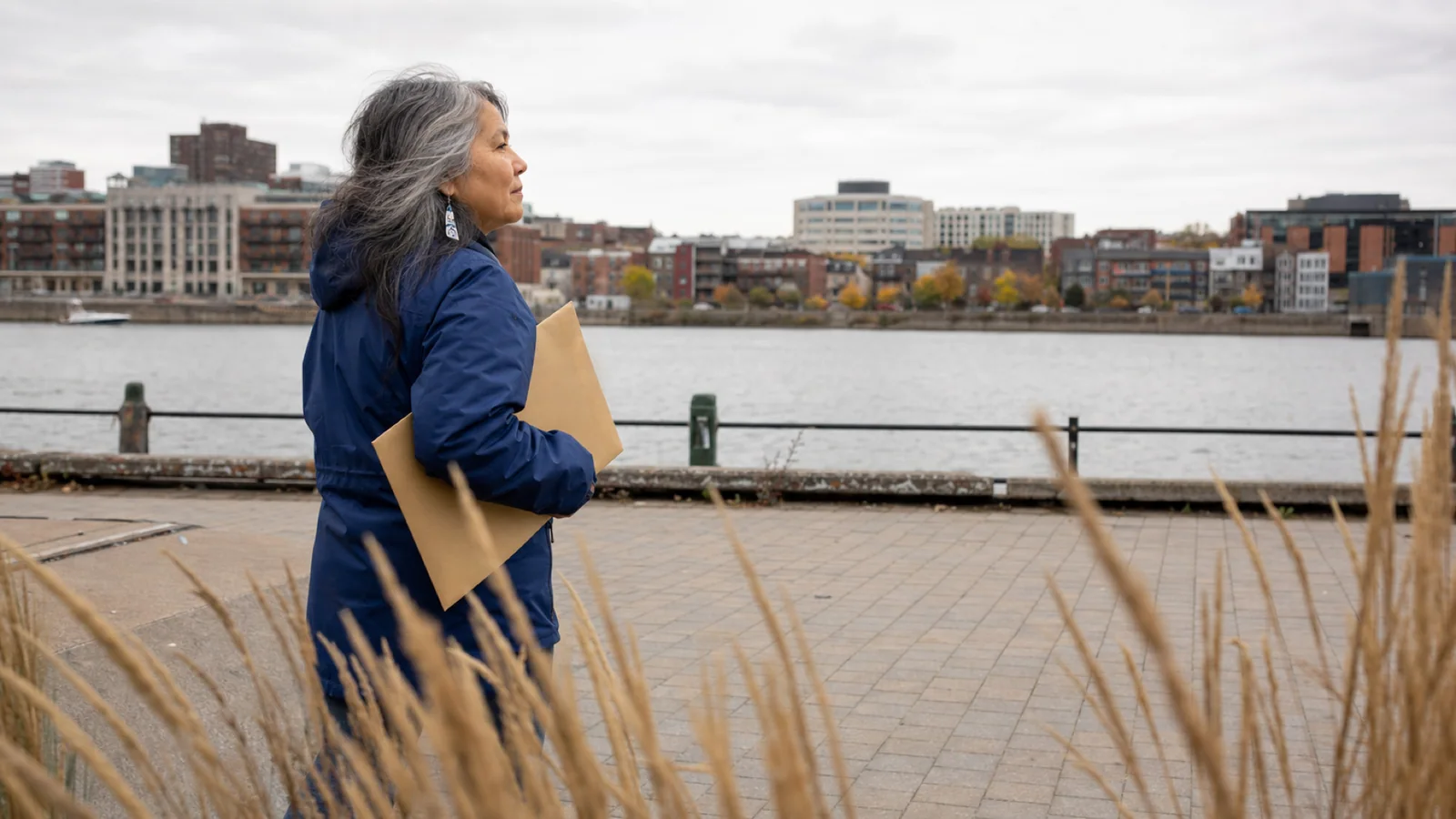 A woman walks along a Quebec river promenade with a closed envelope under her arm.